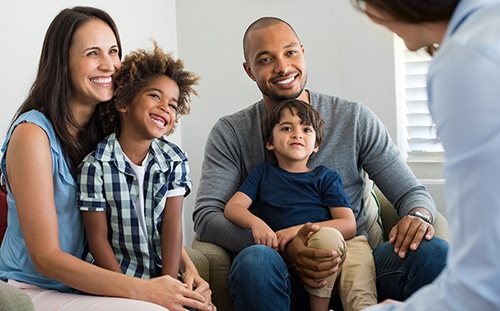 Happy young family sitting on couch and talking with family counselor. Smiling parents with adopted children discussing with counselor. Multiethnic family meeting a financial agent.